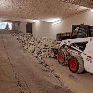 Pozo rectangular excavado, pila de tierra y grava, brazo de maquinaria naranja, árboles en el fondo.