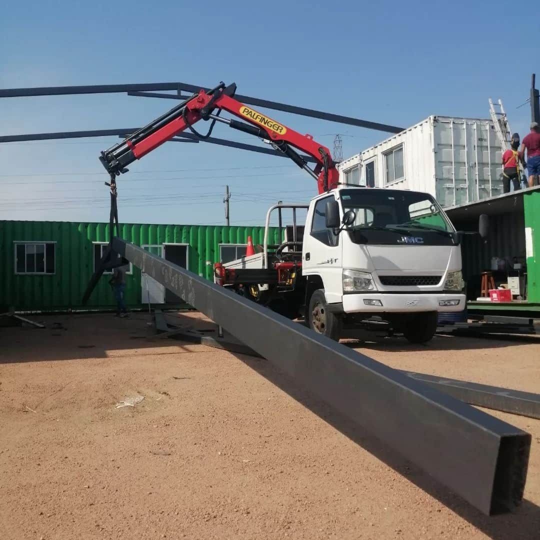 Grúa montada en camión levanta una viga de metal en un sitio de construcción cerca de contenedores verdes y blancos, bajo un cielo soleado.