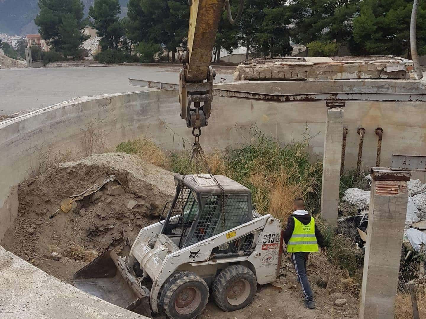 Una minicargadora está sacando un gran bloque de hormigón de una estructura demolida. Una persona con chaleco antibalas está cerca.