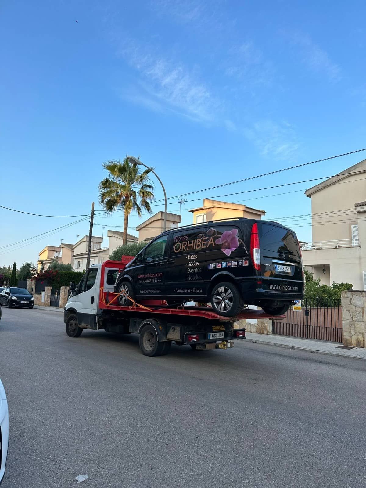 Furgoneta negra sobre una grúa, circulando por una calle bordeada de casas bajo un cielo azul.