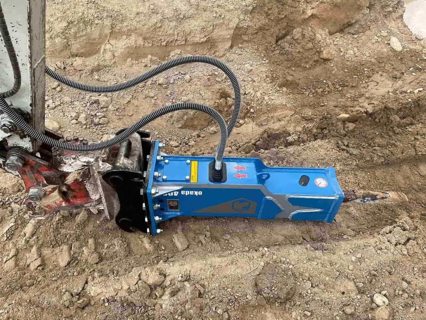 Herramienta rompedora hidráulica azul sobre suelo marrón, conectada a mangueras, rompiendo parcialmente el hormigón.