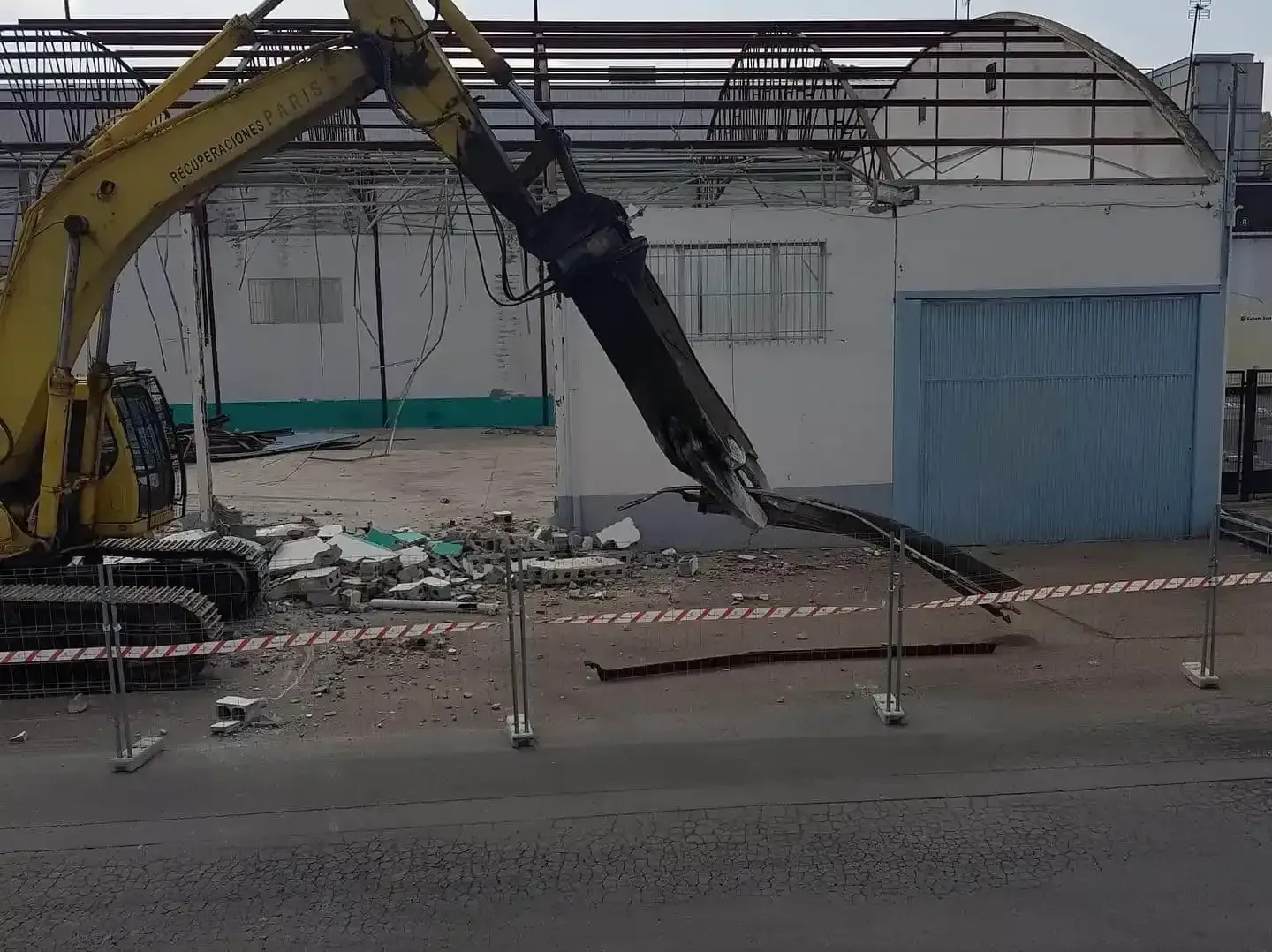 Una excavadora amarilla derriba un edificio blanco con una puerta azul. Escombros y cinta de precaución en primer plano.