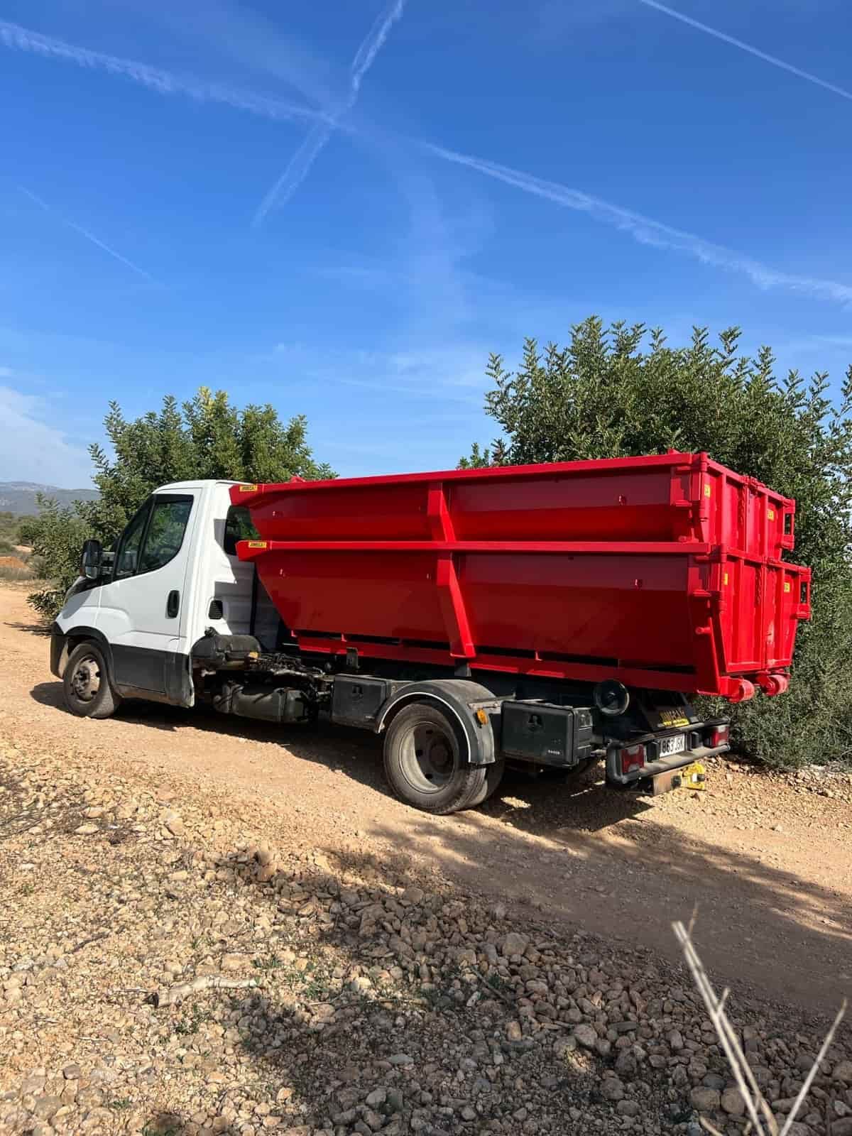 Contenedor rojo en un camión blanco estacionado en un camino de tierra, árboles en el fondo, cielo azul.