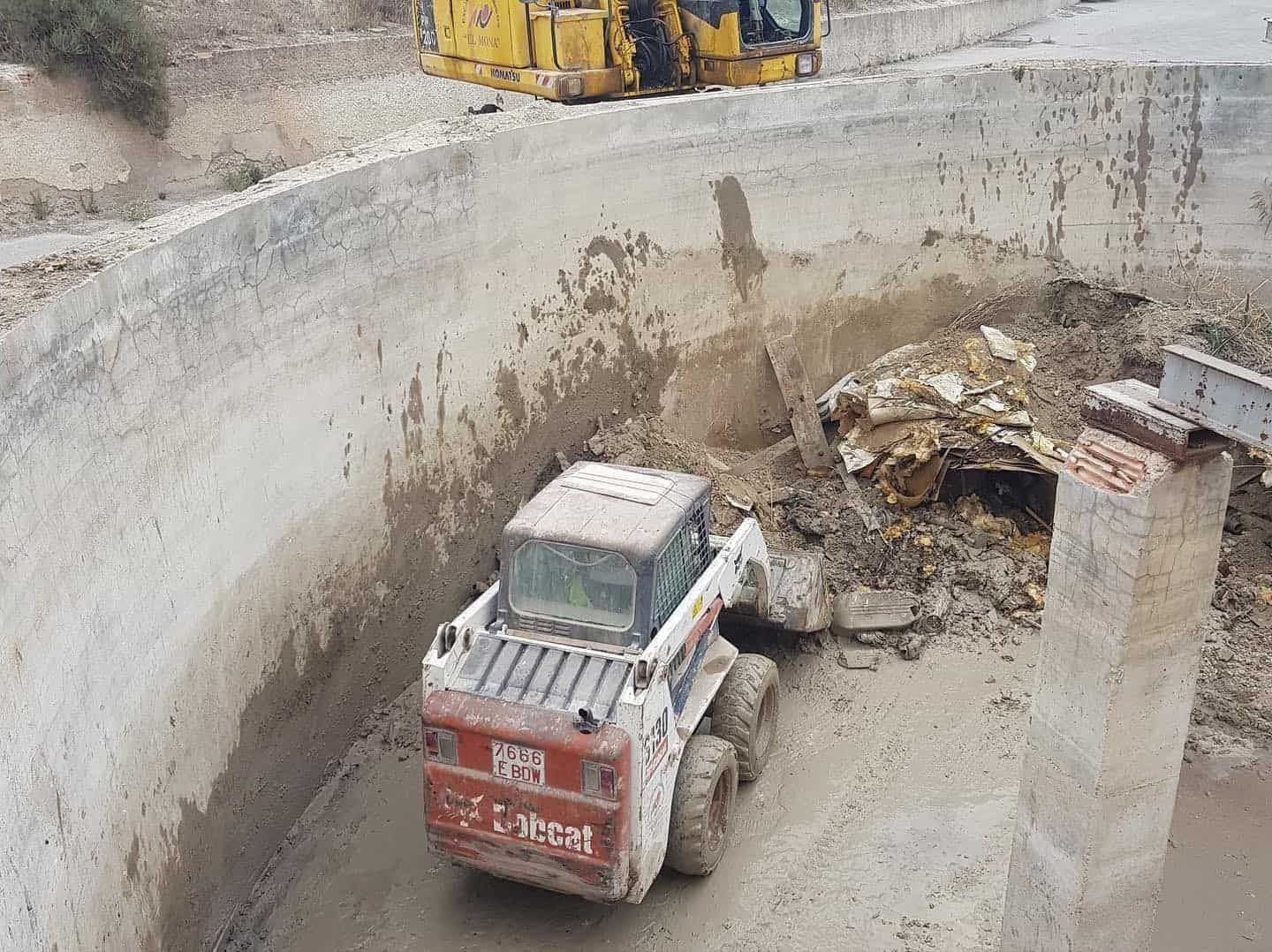 Una minicargadora Bobcat retira escombros de una estructura de hormigón. Se ve una excavadora amarilla al fondo.