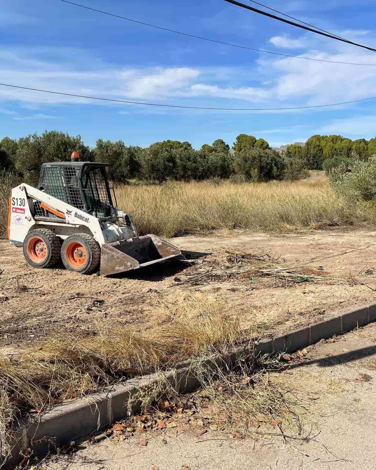 Minicargadora Bobcat desbrozando la maleza. Acera gris, campo marrón y verde, cielo azul.