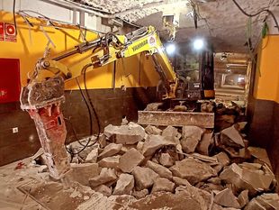 Una minicargadora de color naranja cava una zanja en un patio, observada por un hombre en pantalones cortos y camiseta.