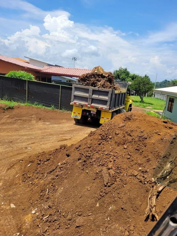 Camión volquete amarillo descargando tierra en un lote de tierra, cerca de una cerca y casas bajo un cielo nublado.