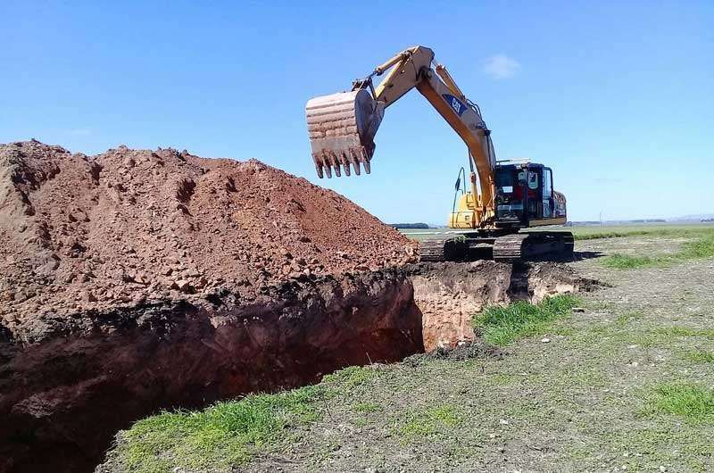 Excavadora amarilla cavando una zanja profunda en un campo de hierba en un día soleado.