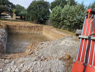 Pozo rectangular excavado, pila de tierra y grava, brazo de maquinaria naranja, árboles en el fondo.