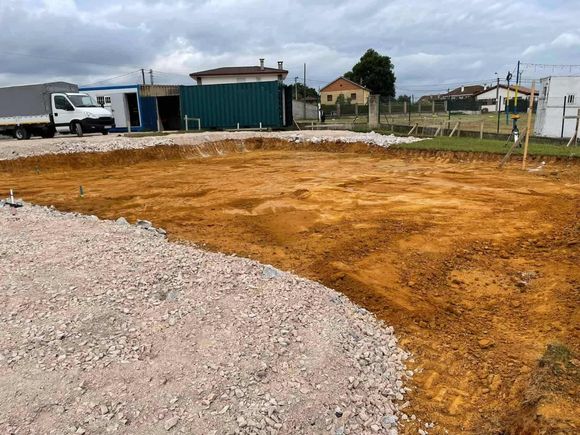 Sitio de construcción: excavación con tierra marrón, camino de grava y vehículos estacionados.
