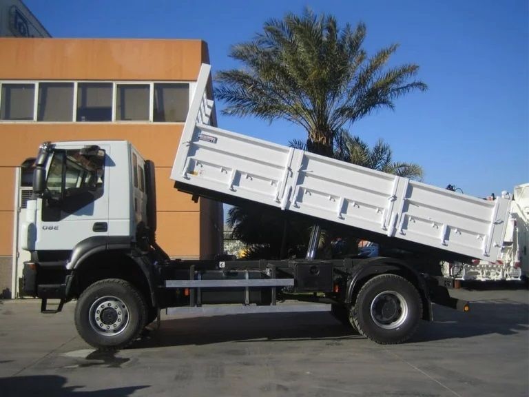 Camión volquete blanco con plataforma elevada frente a un edificio naranja y una palmera.