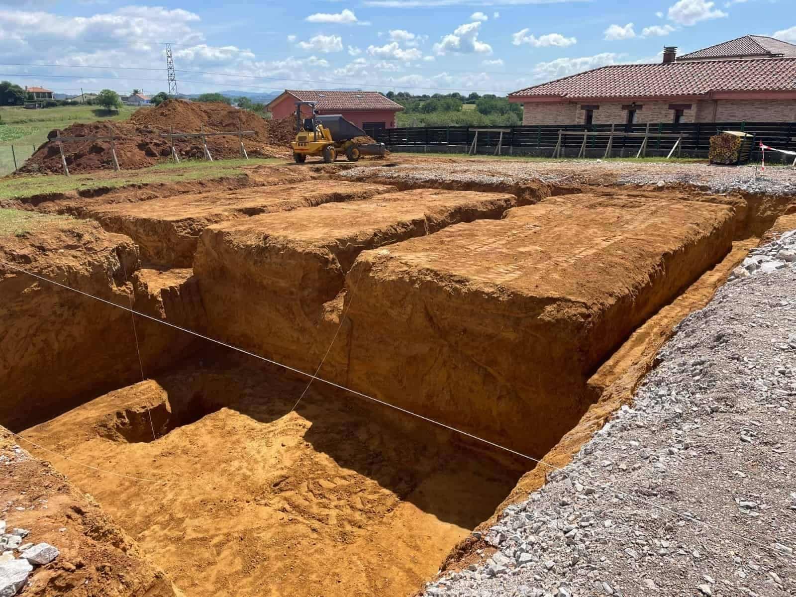 Sitio de excavación para los cimientos de un edificio, con una retroexcavadora visible al fondo en un día soleado.