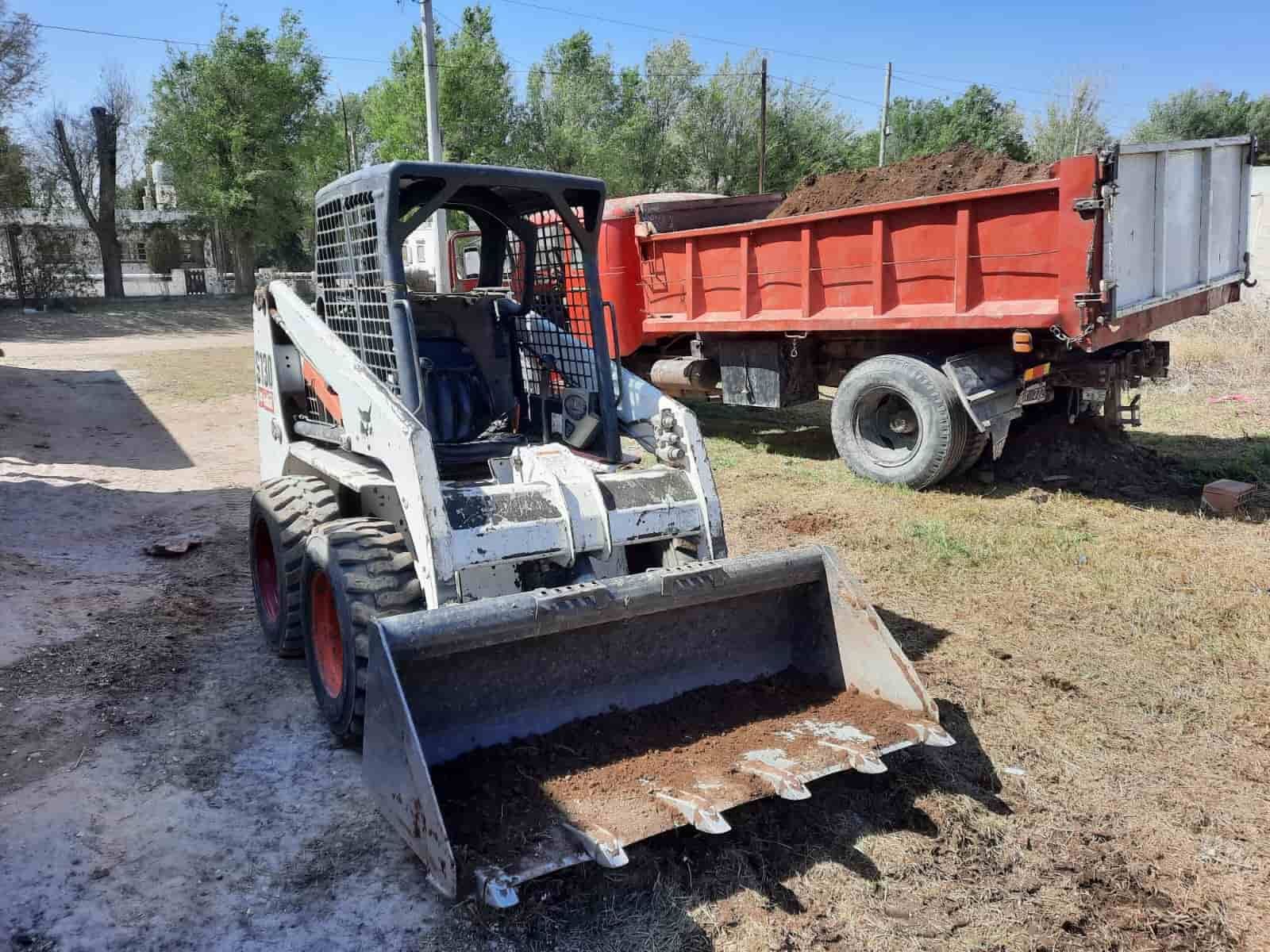 Cargador Bobcat con cucharón al lado de un camión volquete rojo, moviendo tierra en un entorno al aire libre.