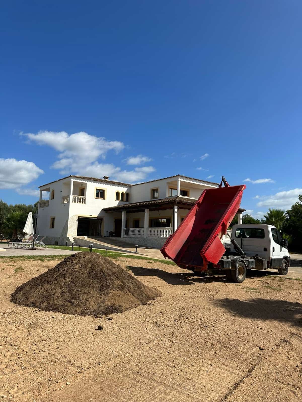 Casa blanca con un camión volquete rojo que arroja tierra en un terreno de tierra bajo un cielo azul.