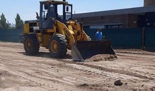 Cargadora amarilla trabajando en un camino de tierra. Una persona está al volante. Edificio al fondo.