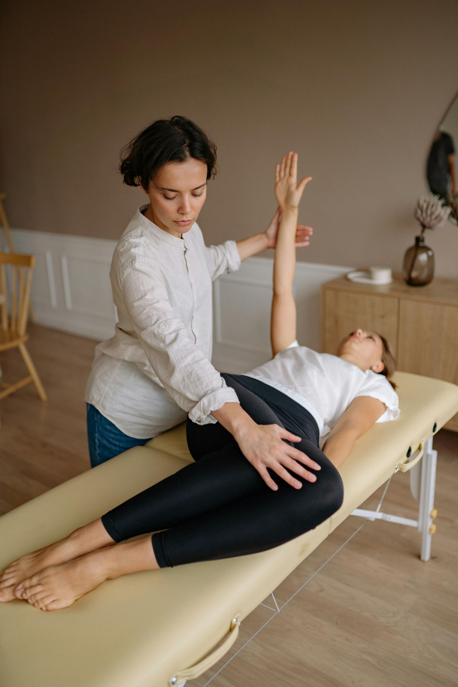 Eine Frau erhält Physiotherapie; der Therapeut stützt Arm und Bein auf einem beigen Tisch in einem Raum.
