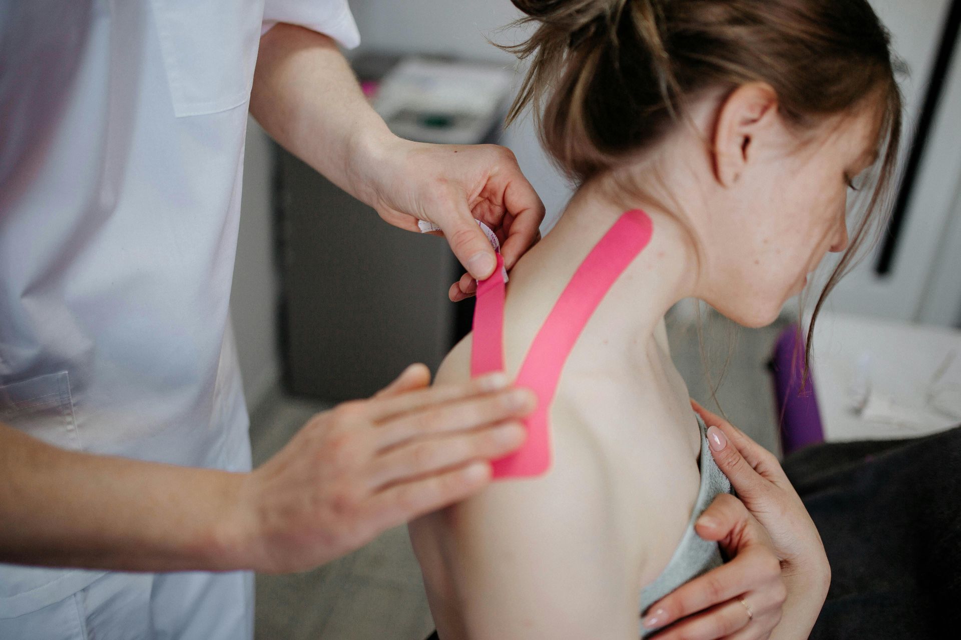 Eine Person klebt in einer medizinischen Einrichtung einer Frau rosa Kinesiologie-Tape an Schulter und Nacken an.