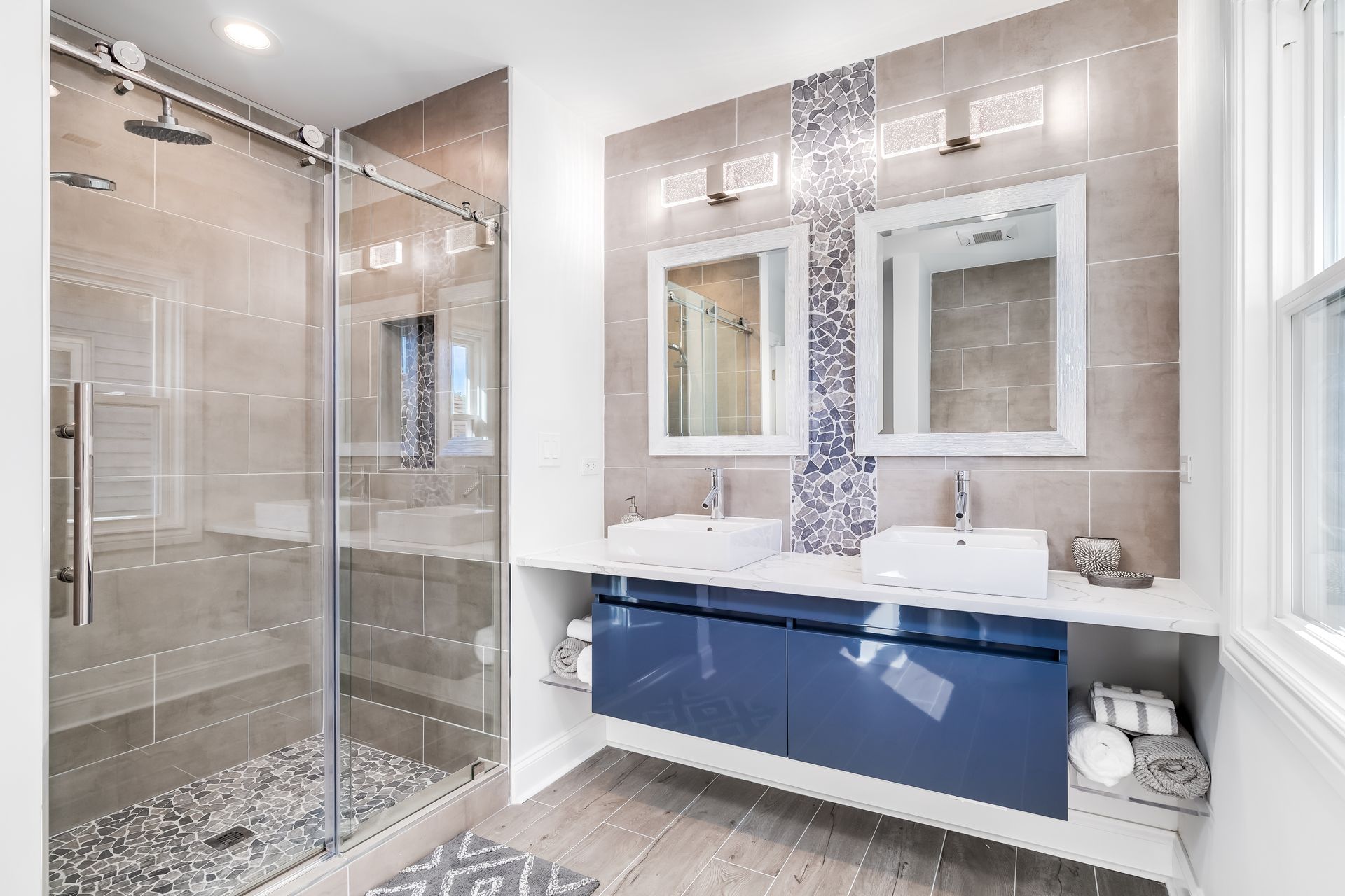 Salle de bains avec double vasque, meuble bleu et douche vitrée. Il y a du carrelage au sol et aux murs.
