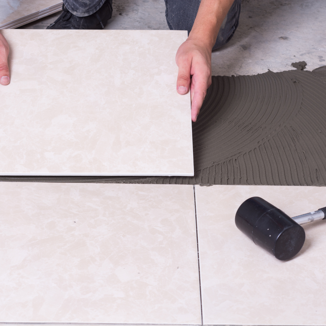 Des mains posent une grande tuile de carrelage sur du mortier gris à côté d'une autre tuile.