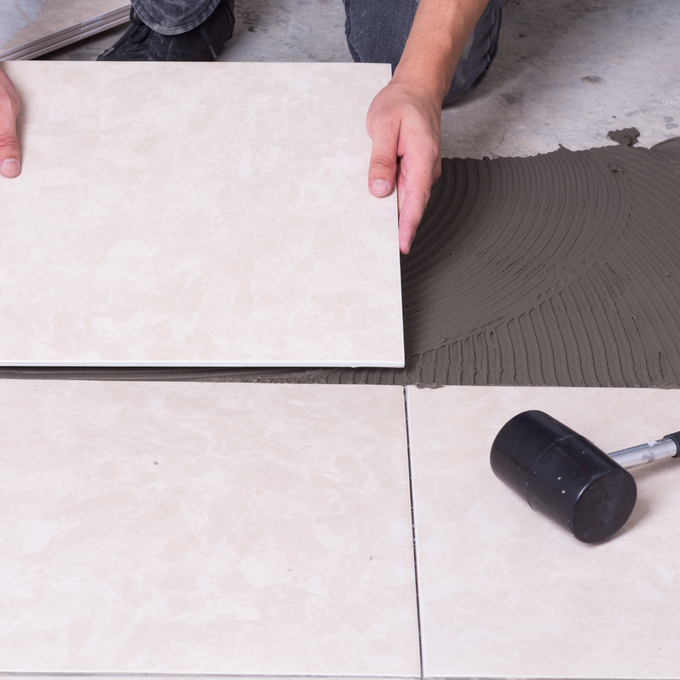 Des mains posent une grande tuile de carrelage sur du mortier gris à côté d'une autre tuile.