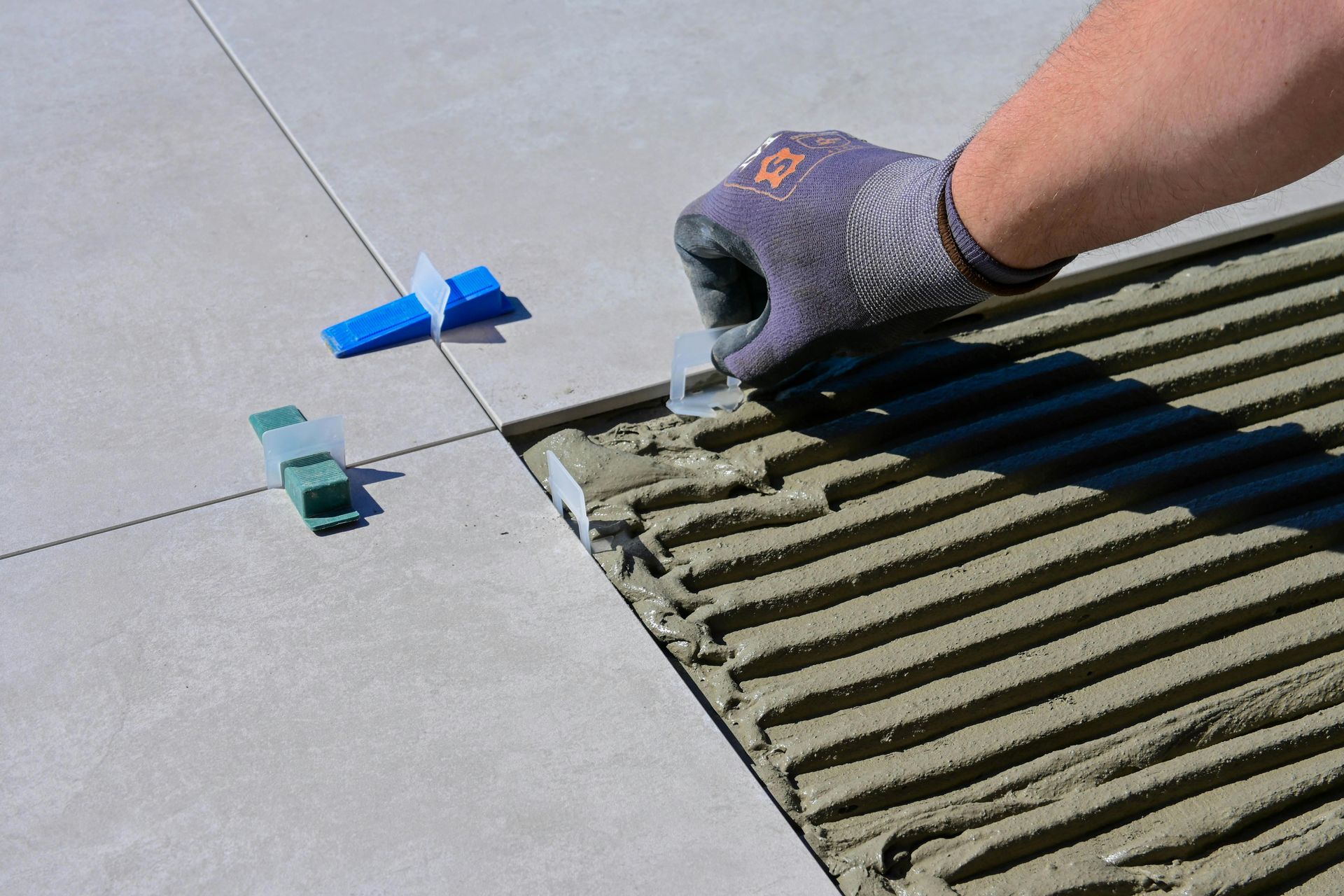 Une main gantée étale de la colle à carrelage grise sur un sol partiellement carrelé.
