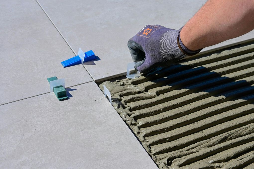 Une main gantée étale de la colle à carrelage grise sur un sol partiellement carrelé.