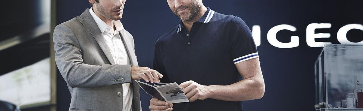 Deux hommes regardent une tablette, l'un en blazer, l'autre en polo, avec le logo Peugeot en arrière-plan.