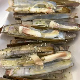 Montón de navajas crudas sobre un plato blanco, con las conchas abiertas dejando ver la carne y las hierbas.
