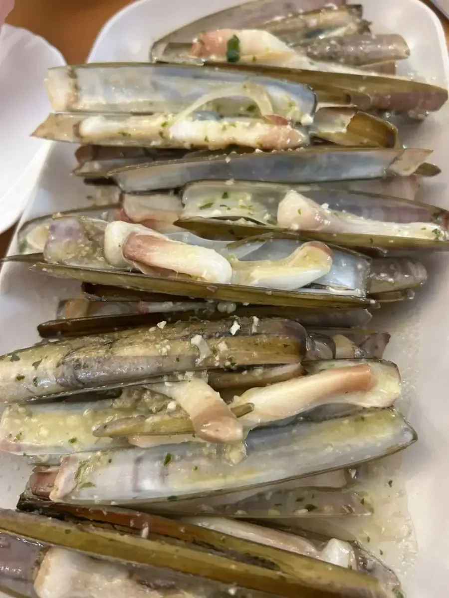 Montón de navajas crudas espolvoreadas con condimentos sobre un plato blanco.