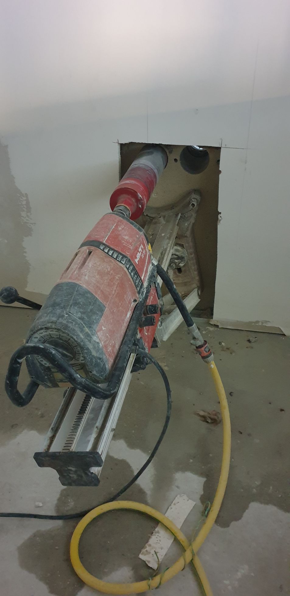 Une carotteuse fixée au mur est en train de le percer. Un tuyau d'arrosage jaune est branché et le sol est mouillé.