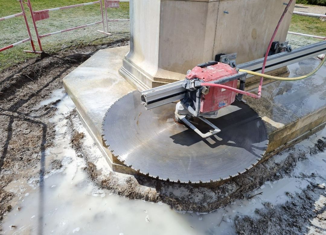 Une grande scie circulaire découpe du béton autour de la base d'une structure en pierre.