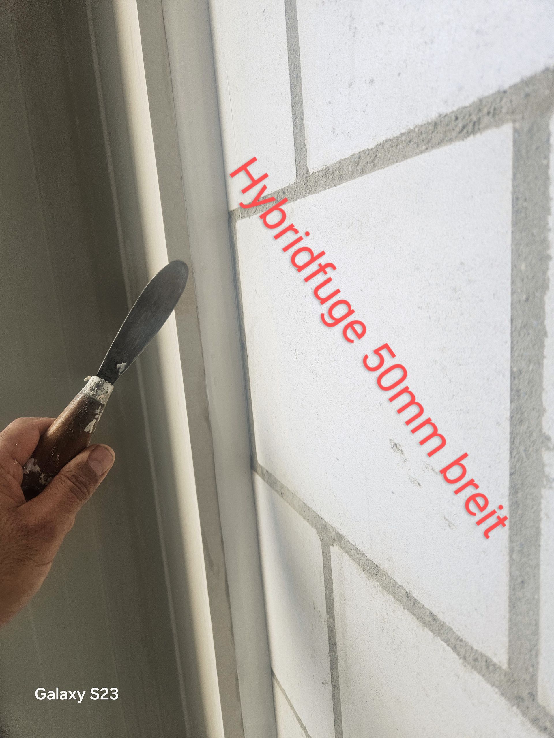 Eine Person hält einen Spachtel vor einer Wand mit der Aufschrift „Hybridfuge 50mm breit“
