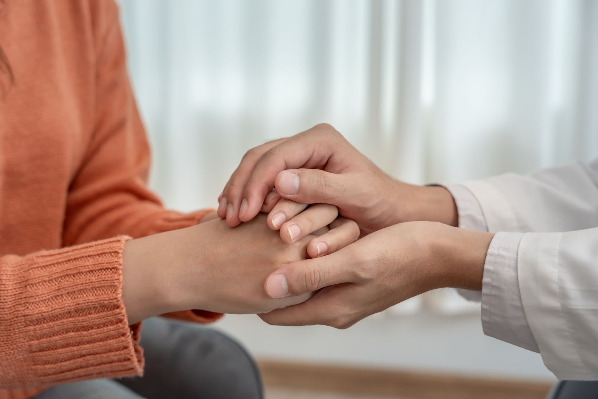 Les mains d'une personne sont jointes en signe de réconfort par une autre personne en blouse blanche, à l'intérieur.