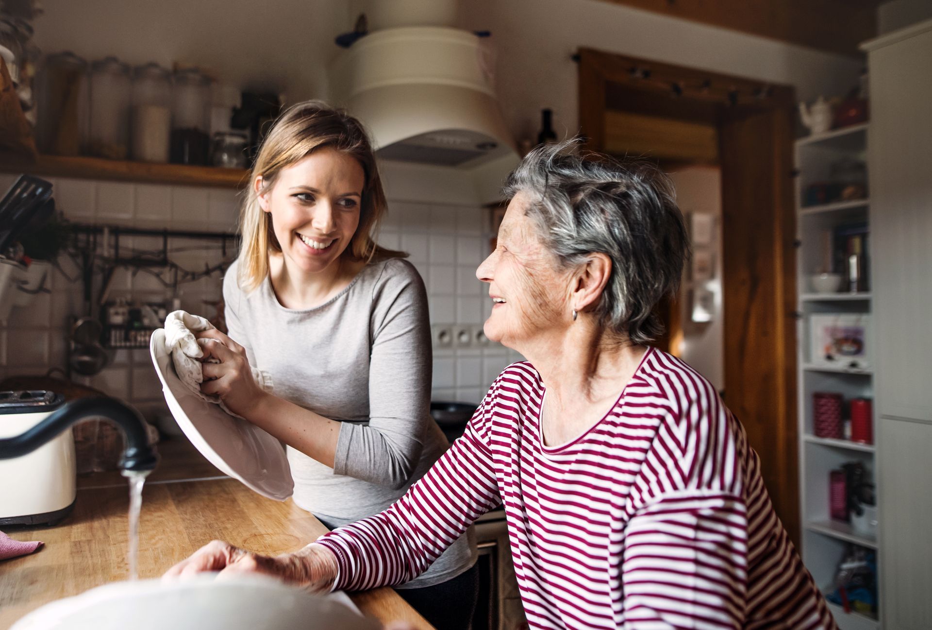 Une femme fait la vaisselle avec une femme âgée dans une cuisine, toutes deux souriantes.