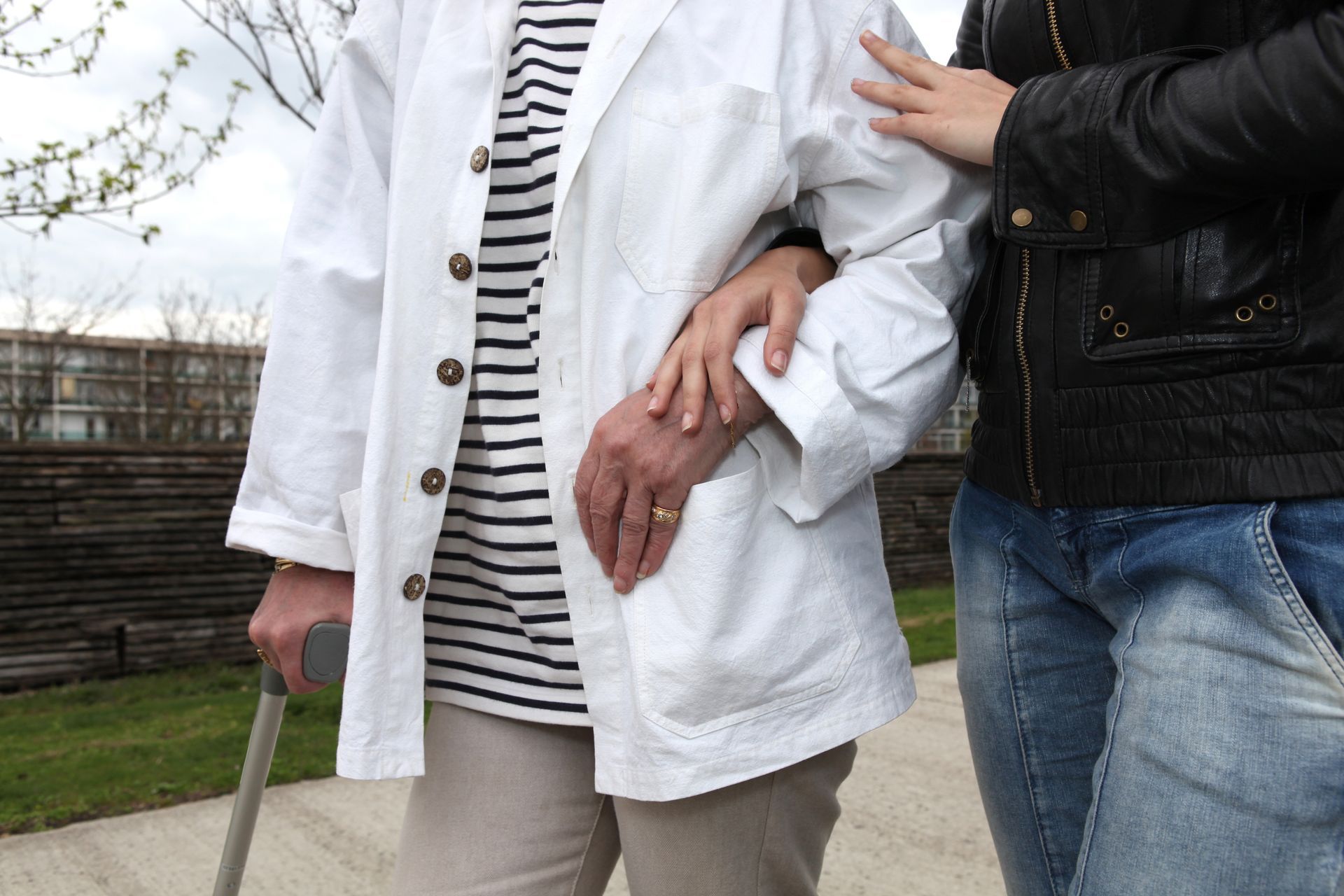 Personne avec canne, vêtue d'une blouse blanche et d'une chemise rayée, assistée par une autre personne à l'extérieur.