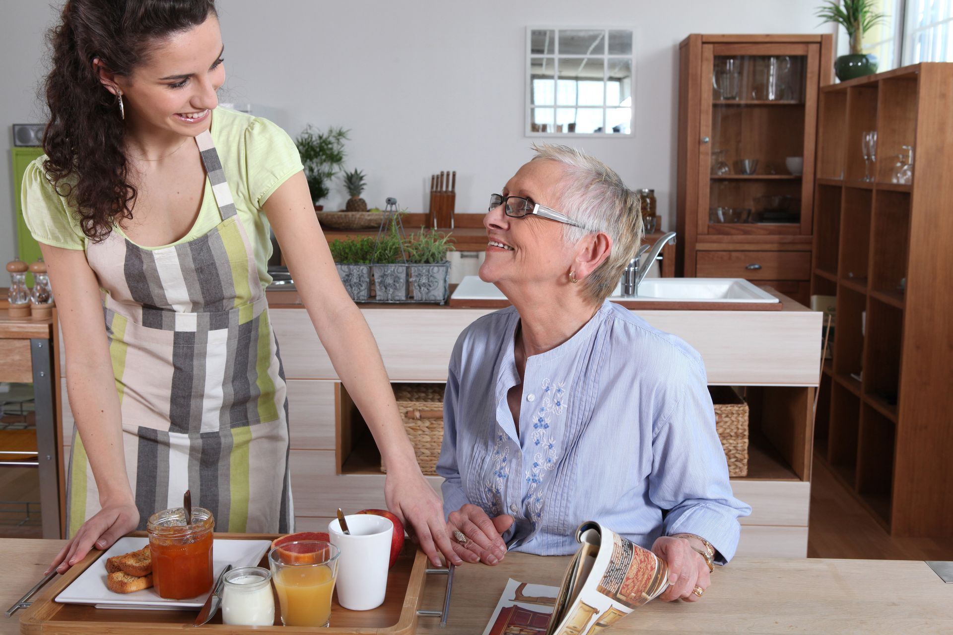 Une jeune femme sert le petit-déjeuner à une femme âgée dans une cuisine, en souriant.