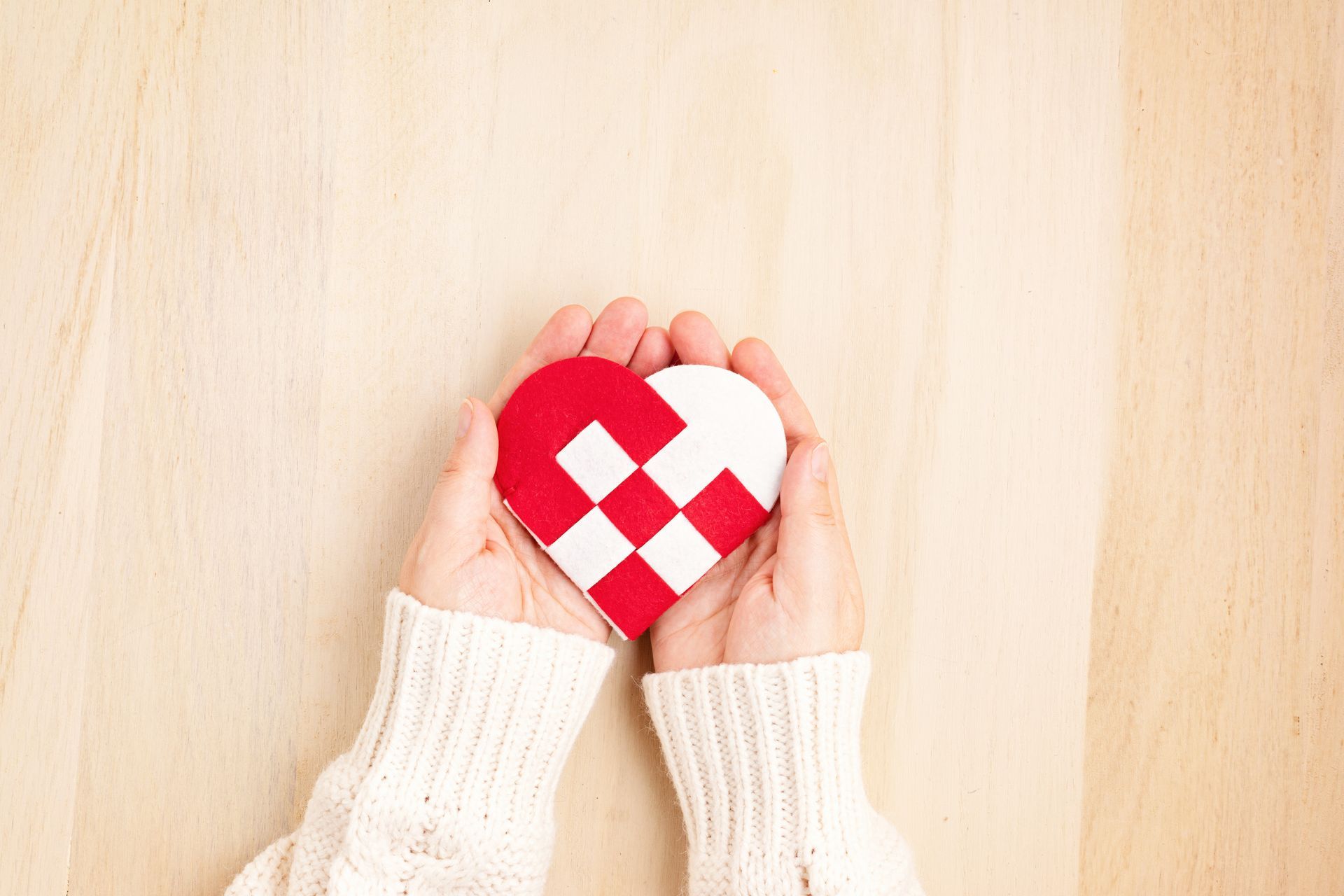Mains tenant un ornement en forme de cœur avec un motif tissé rouge et blanc sur un fond en bois clair.