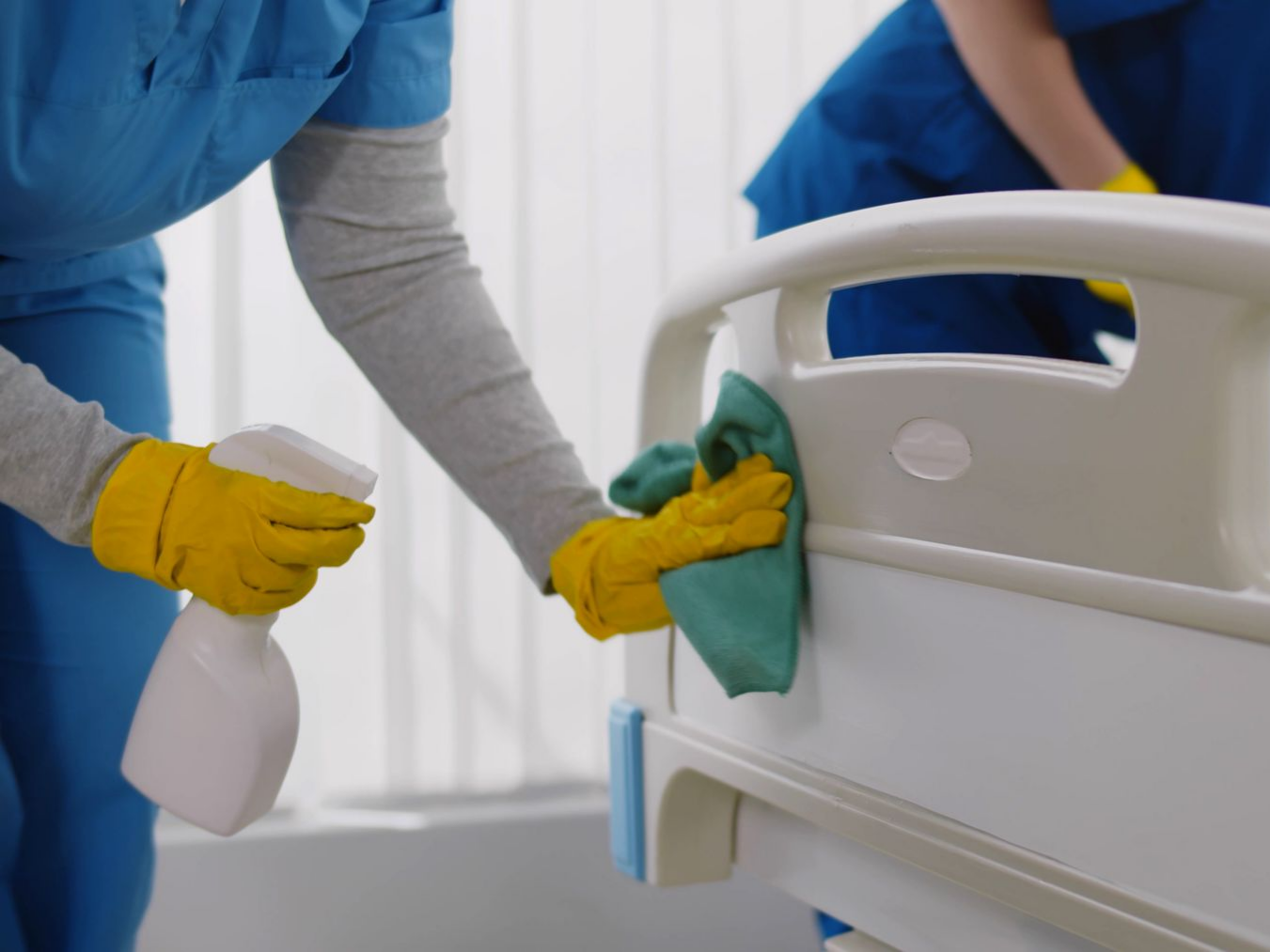 Un agent de nettoyage nettoie la barrière d'un lit d'hôpital avec un chiffon et un vaporisateur.