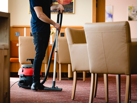 Une personne passe l'aspirateur dans une pièce recouverte de moquette, avec des chaises beiges et des meubles en bois.
