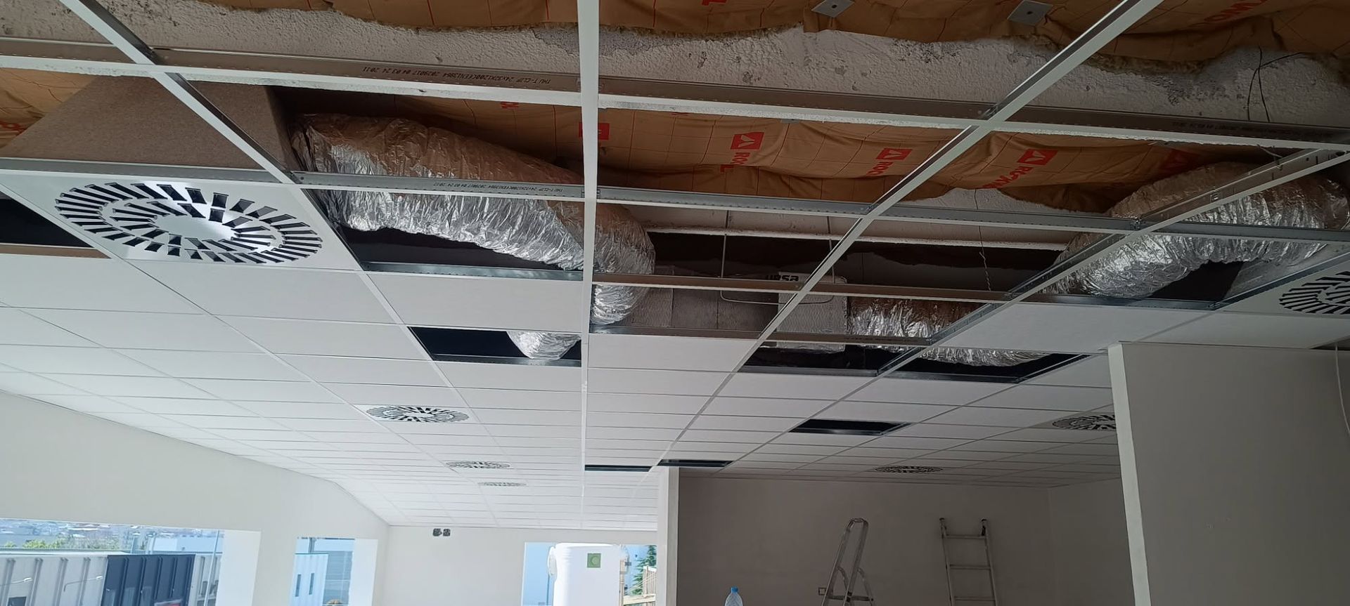 Techo de oficina dañado, con placas faltantes, aislamiento expuesto y luminarias colgantes durante la renovación