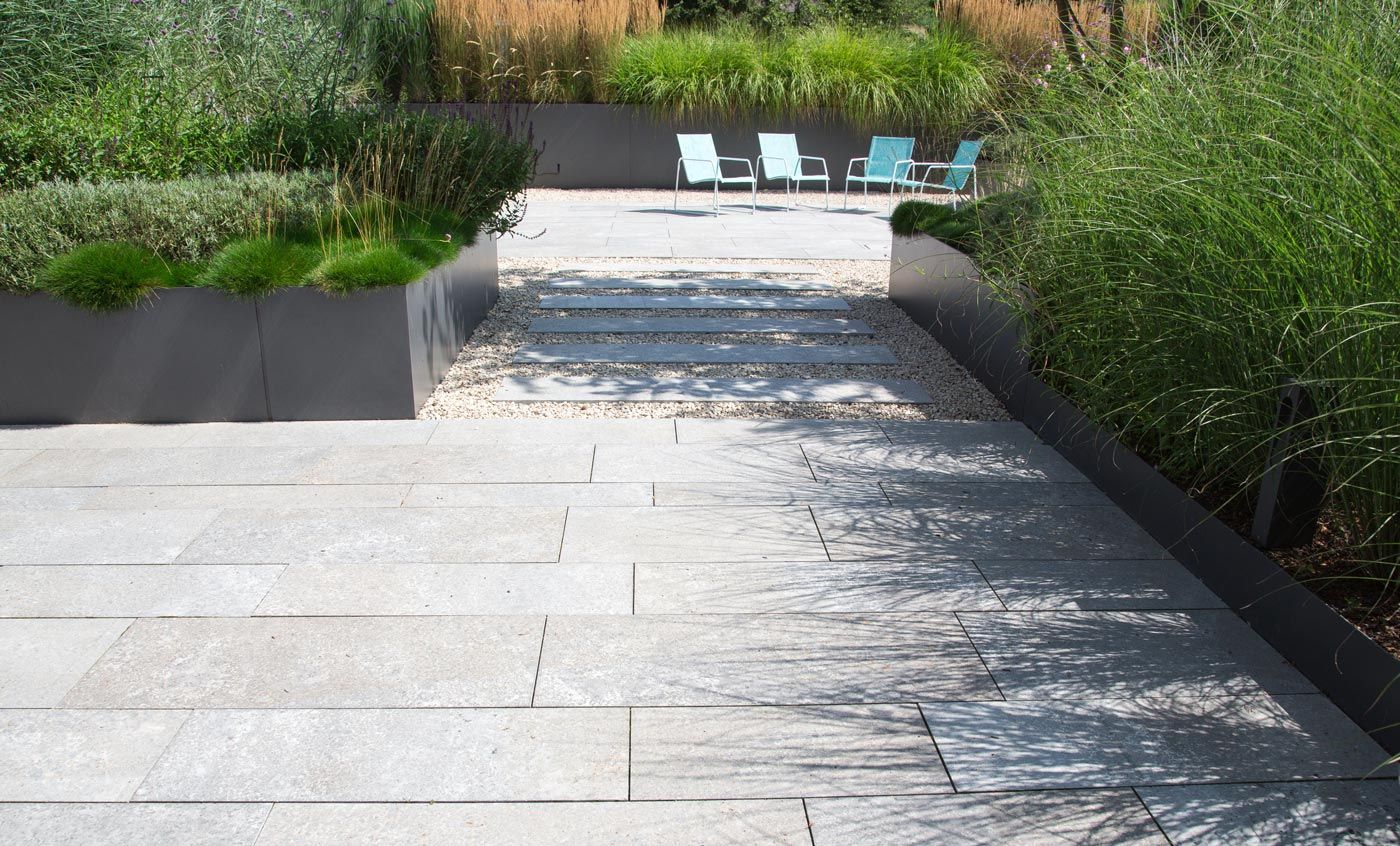 Une terrasse de jardin en carrelage de pierre, des parterres de plantes vertes autour