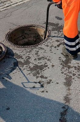Un trabajador con equipo de seguridad naranja limpia una alcantarilla en una calle mojada.