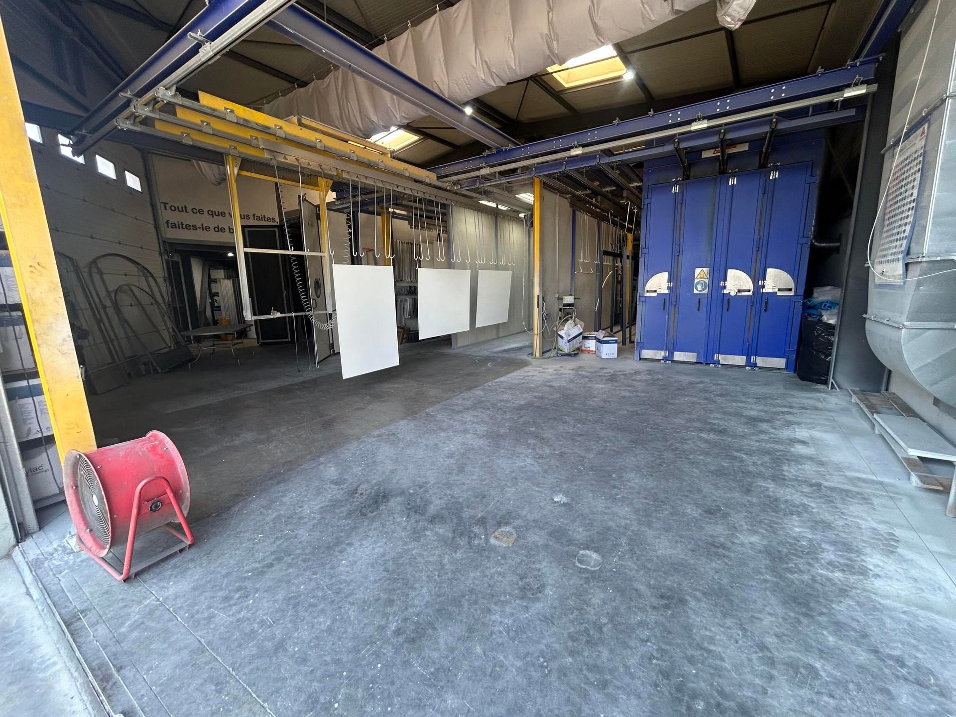 Atelier industriel vide avec sol en béton, poutres métalliques, rideau bleu et ventilateur rouge au premier plan