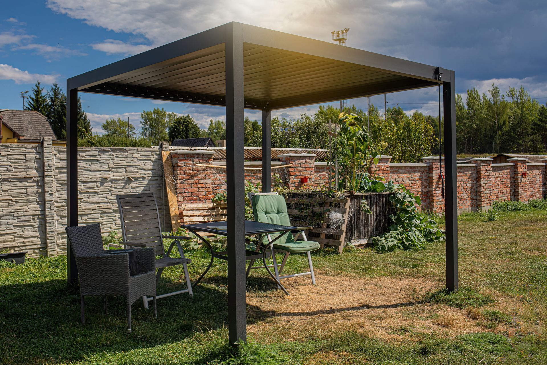 Pergola en métal avec des chaises et une table, dans un jardin herbeux près d’un mur en pierre.