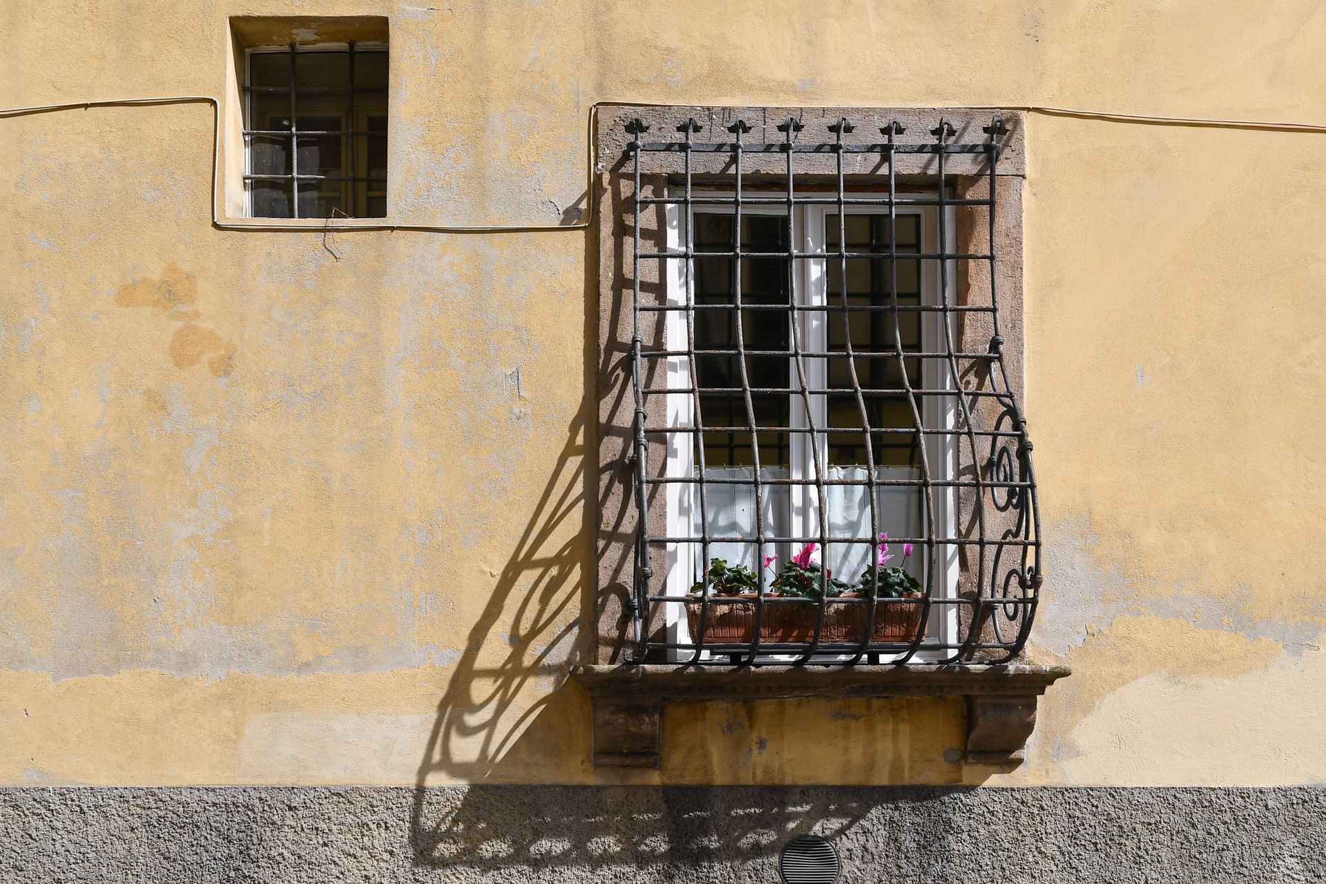 Petite fenêtre à barreaux avec jardinière sur un mur en stuc jaune, projetant une ombre au soleil
