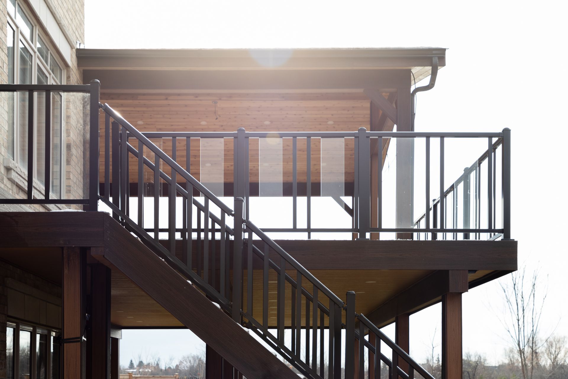 Terrasse extérieure en bois avec rambardes et escaliers noirs, jouxtant une maison en briques baignée de soleil.