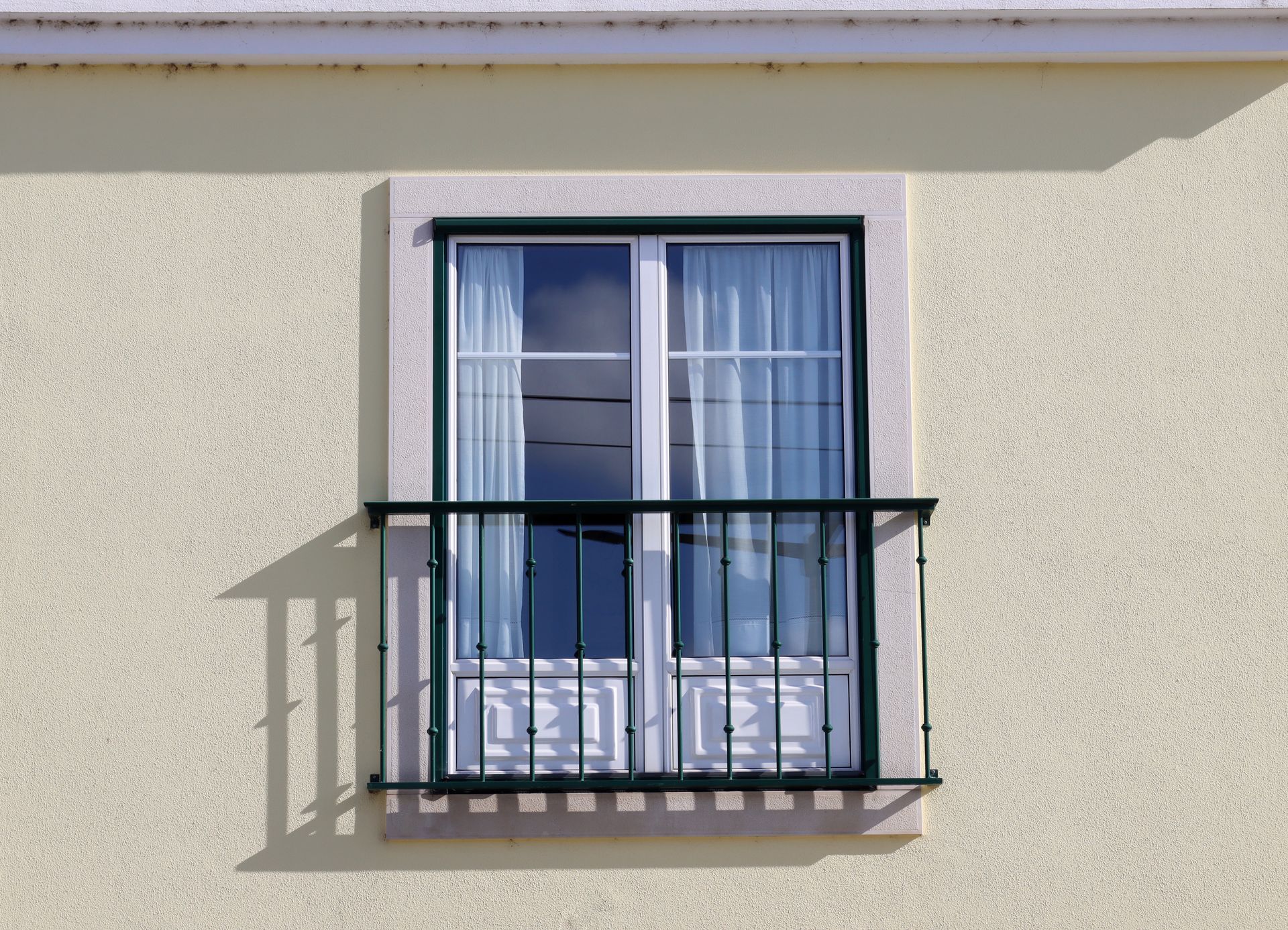 Balcon avec rambarde noire devant un mur en stuc crème et une fenêtre à cadre bleu avec rideaux.