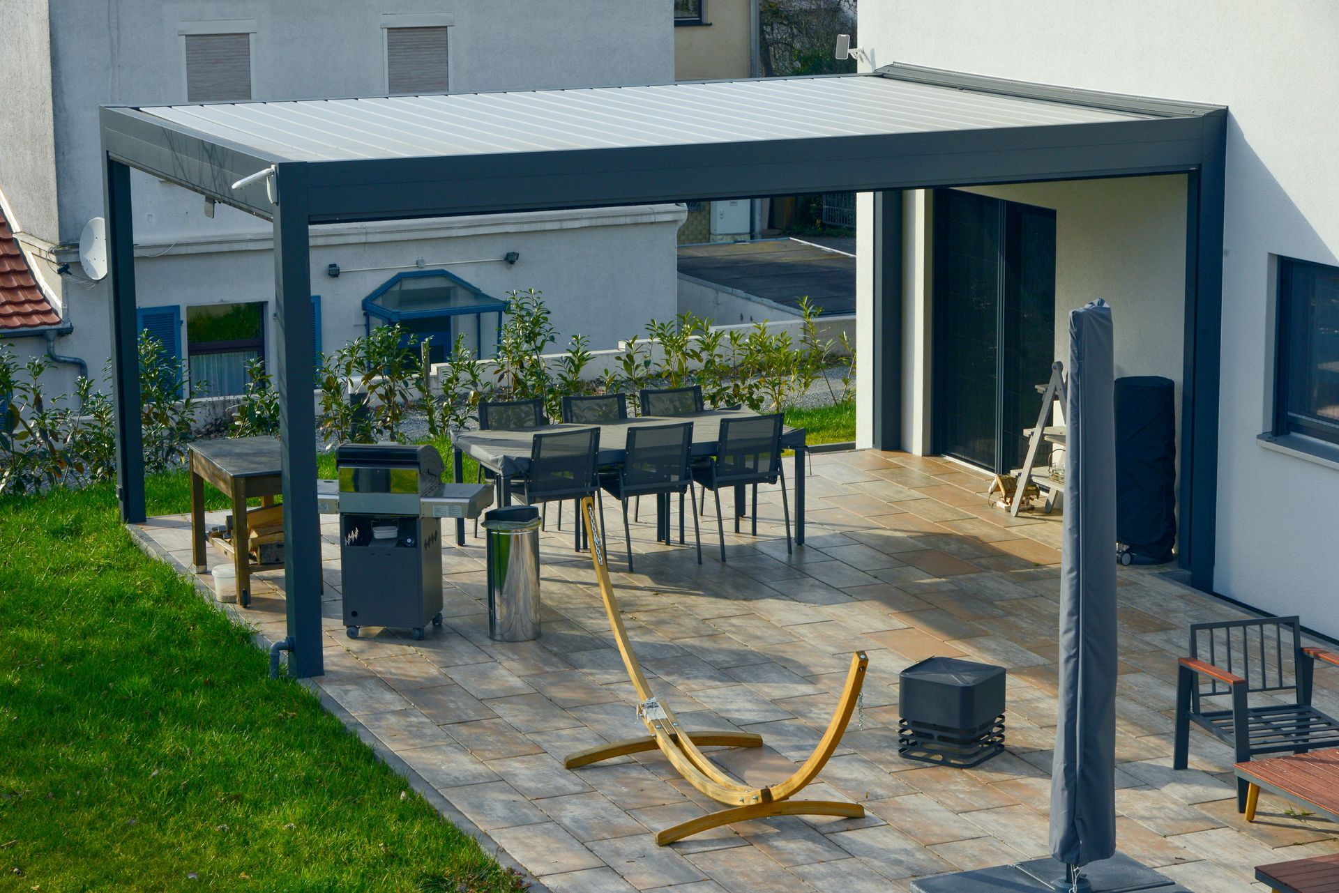 Terrasse avec pergola, table et chaises de jardin, tuyau d'arrosage sur dalles de pierre à côté d'une maison
