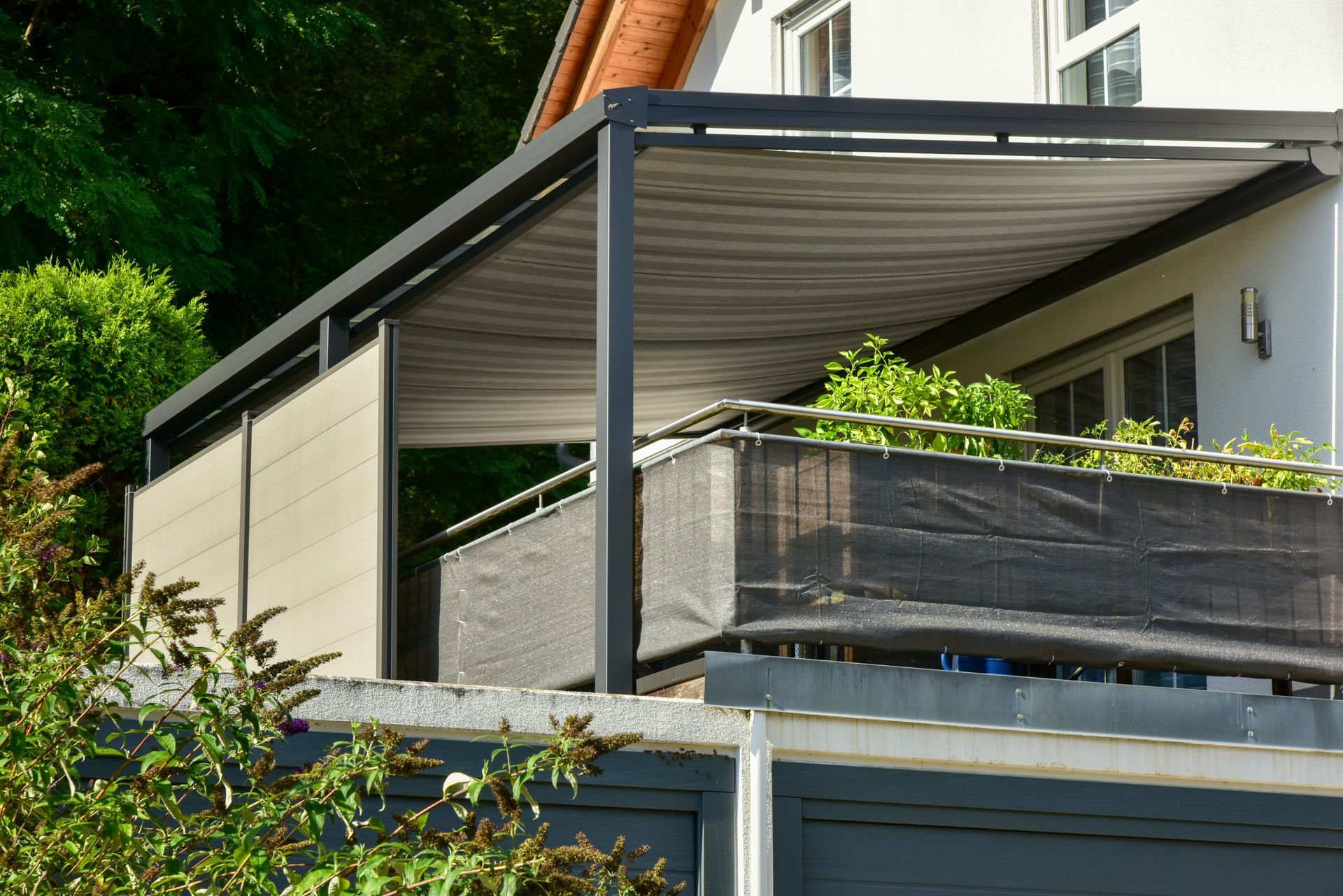 Terrasse couverte avec auvent métallique et rambarde en treillis noir, jouxtant une maison et entourée de verdure.