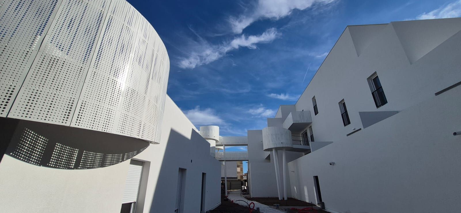 Des immeubles modernes blancs bordent une ruelle étroite sous un ciel bleu parsemé de nuages ​​épars.